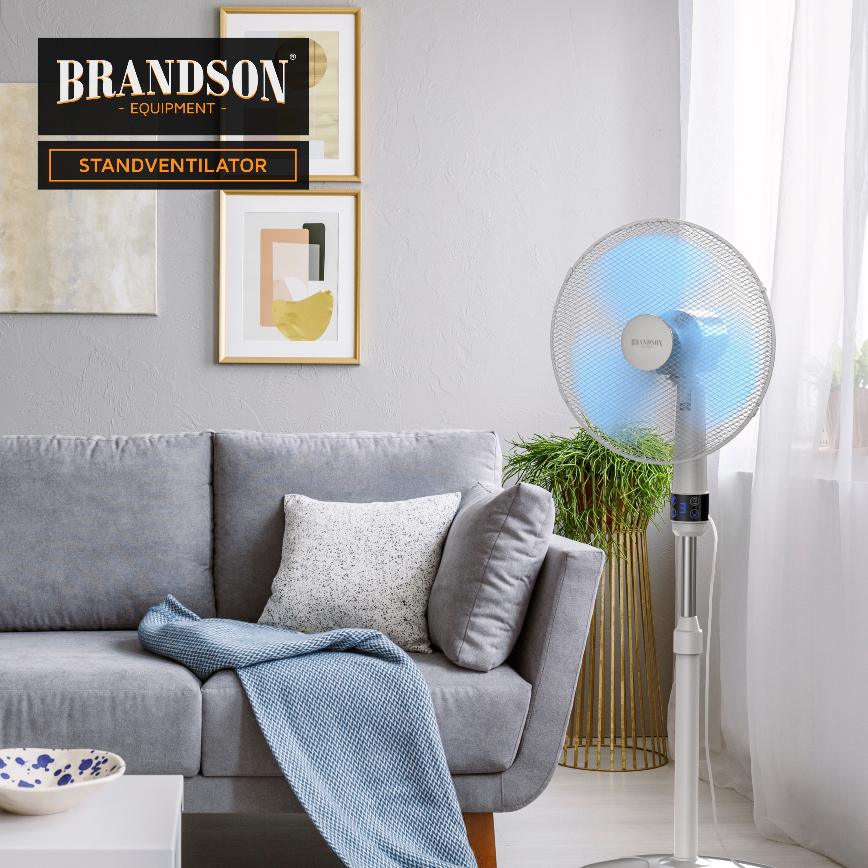 Brandson Standventilator im modernen Design mit silbernem Gehäuse und transparentem Schutzgitter, platziert neben einem Sofa.