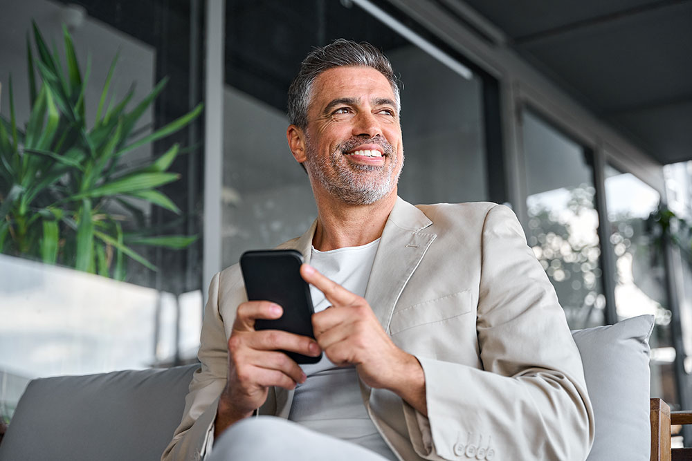 Ein lächelnder Mann in einem hellen Anzug sitzt auf einer Couch und hält ein Smartphone in der Hand, umgeben von viel Grün in einem modernen Ambiente.