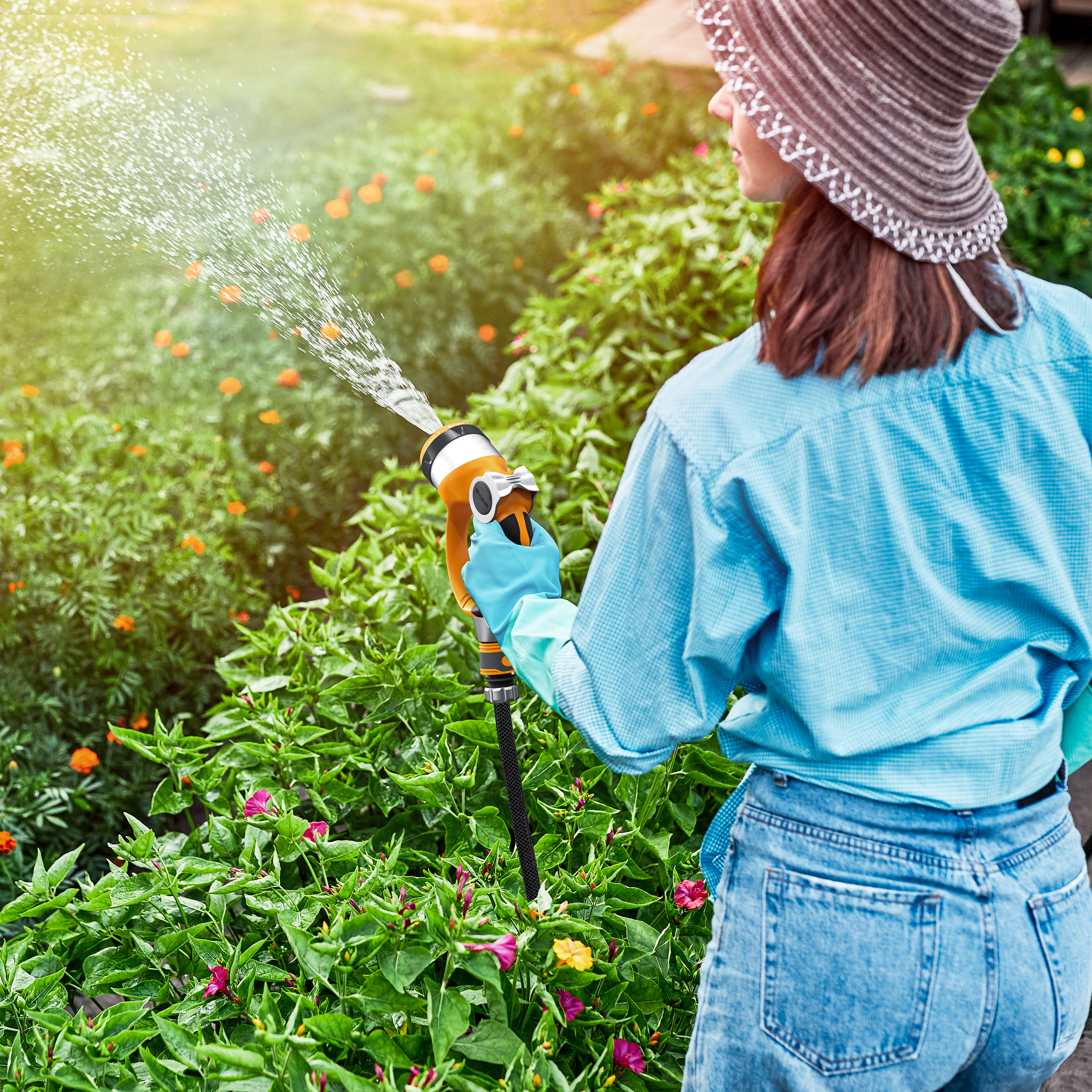 Eine Frau gießt Blumen in einem Garten mit einer Handsprühdüse mit orangefarbenem Griff und blauen Handschuhen und trägt einen Sonnenhut.