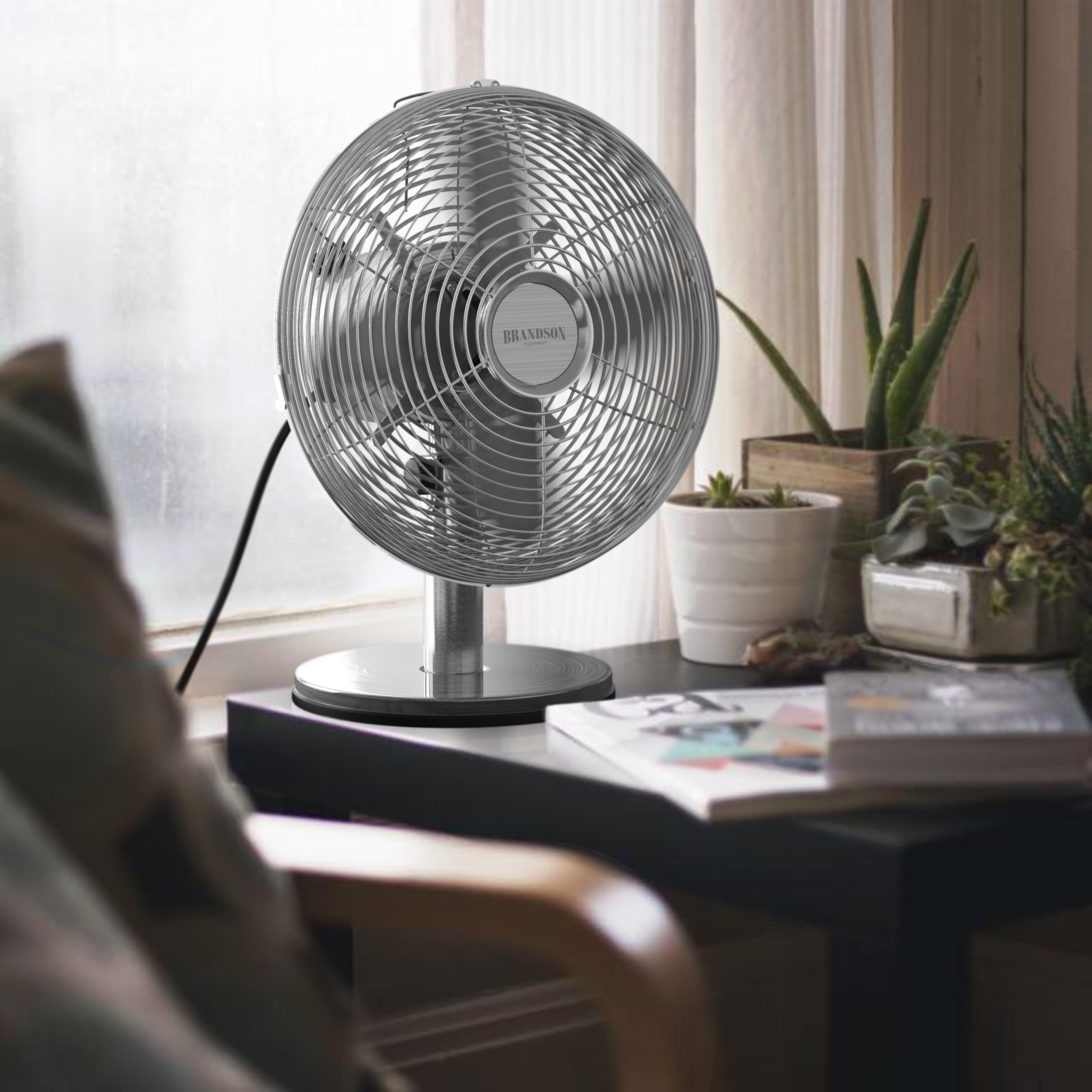 Standventilator von Brandson mit elegantem silbernen Design, Metallgitter und stabilem Sockel, aufgestellt auf einem Tisch in der Nähe von Pflanzen und Büchern.