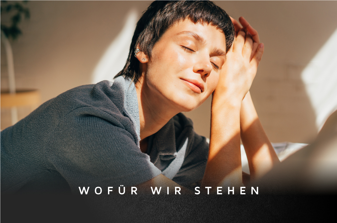 Eine Frau mit kurzen Haaren entspannt sich mit geschlossenen Augen und gefalteten Händen in einem sonnendurchfluteten Raum mit sanften Schatten.